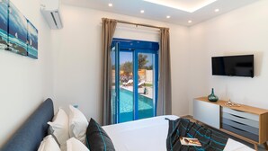 Ein Schlafzimmer mit Bett, Fernseher, Nachttisch, einem Fenster mit Vorhängen und Blick auf einen Außenpool.