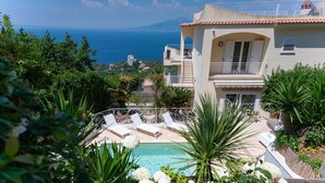 A pool area with lounge chairs, surrounded by lush greenery and a view of the sea.
