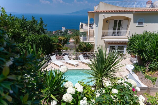 Zona de piscina con sillones de descanso, rodeada de abundante vegetación y vistas al mar.