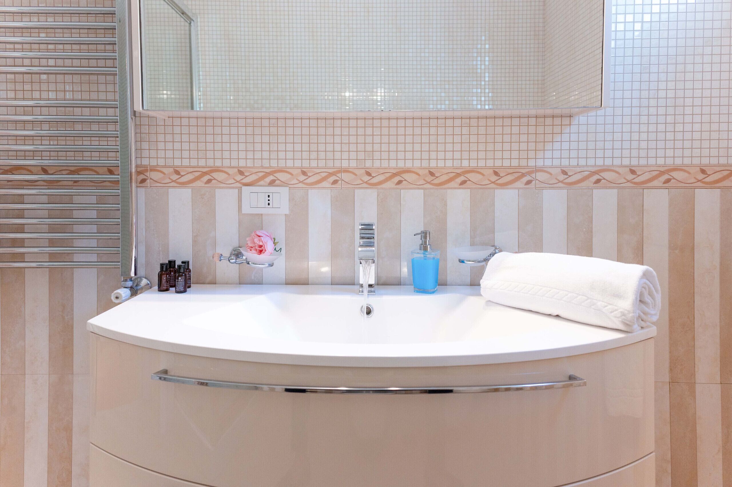 A bathroom with a large oval sink, a mirror above it, and a towel on the right. There are toiletries on the sink and a decorative tile border.