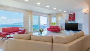 A modern living room with a large sofa, a red armchair, and a television. There is a large window with a view of the sea and a mountain.