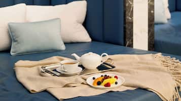 A neatly arranged bed with a blue headboard, a bedside table with a teapot, a cup, a plate of fruit, and a blanket.