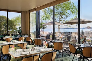 An outdoor dining area with tables and chairs, large windows, and umbrellas overlooking a body of water. - The Woodward, Auberge Collection (Geneva)
