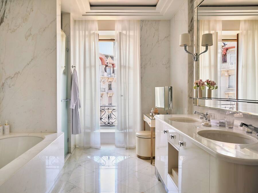 A luxurious bathroom with a large marble sink, a bathtub, and a glass-enclosed balcony with a view of buildings.