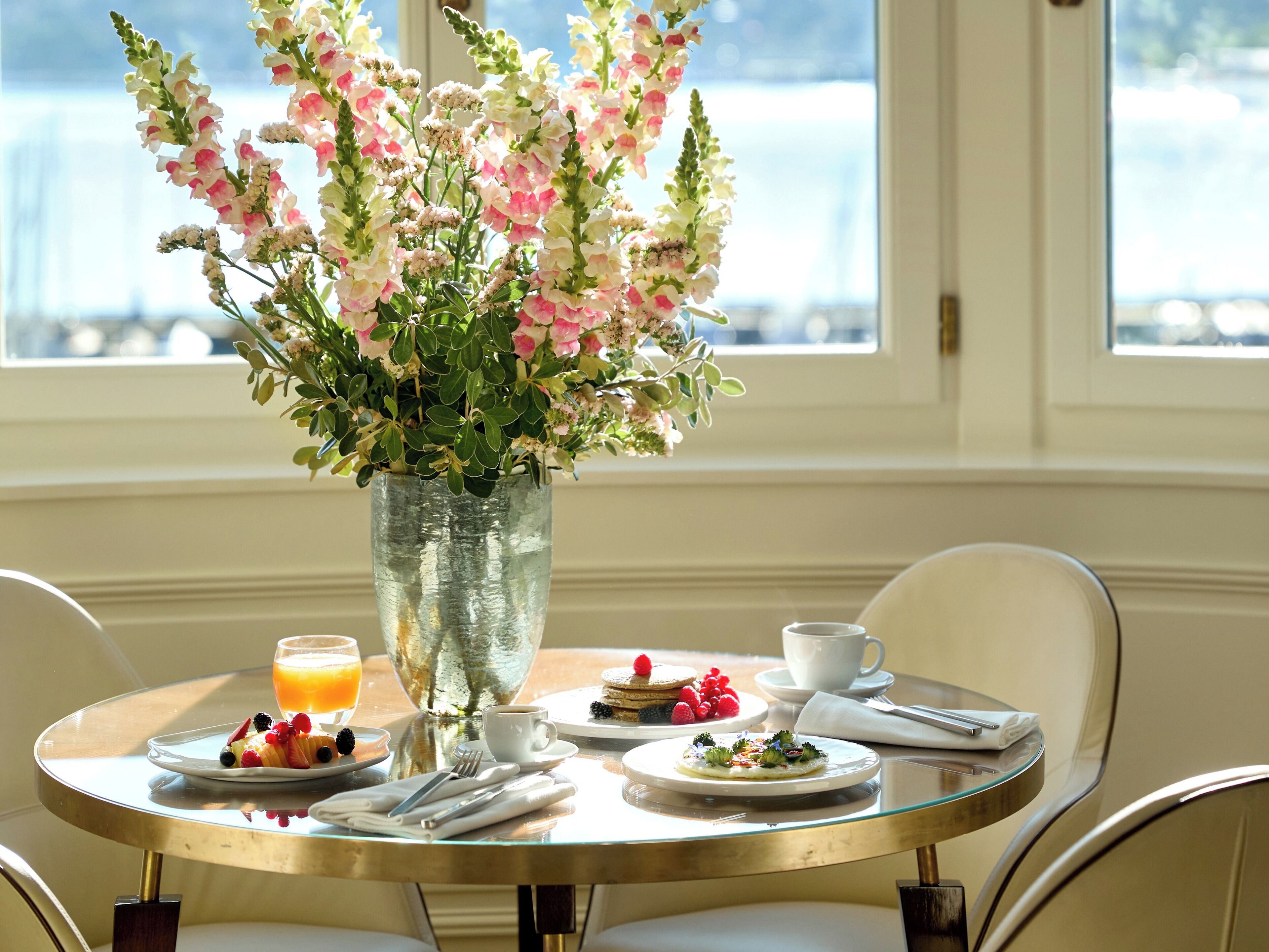 A dining table with a floral arrangement, breakfast items, and a view of the outdoors.