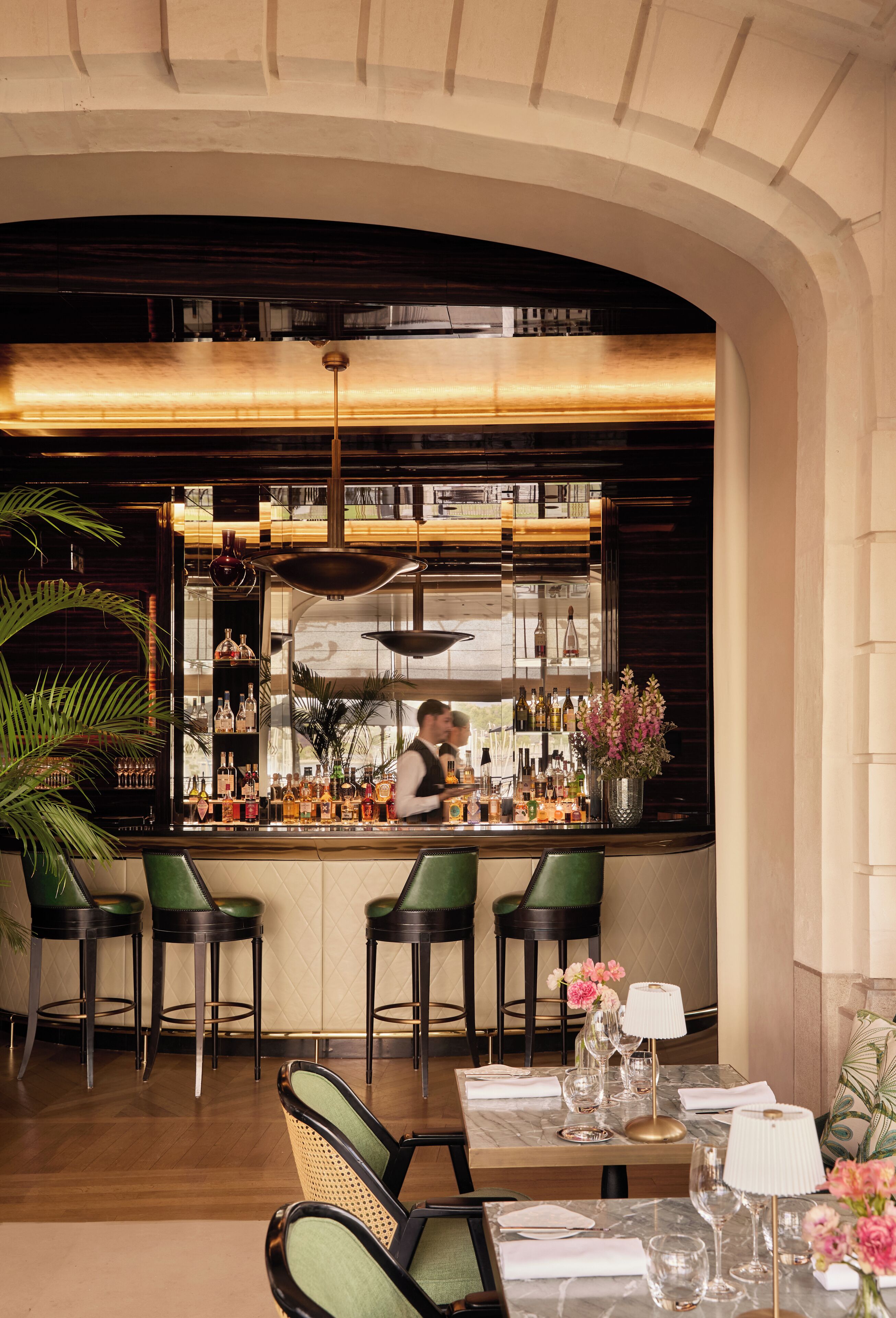 A bar area with a bartender, green bar stools, a marble-topped table with glasses, and a potted plant.