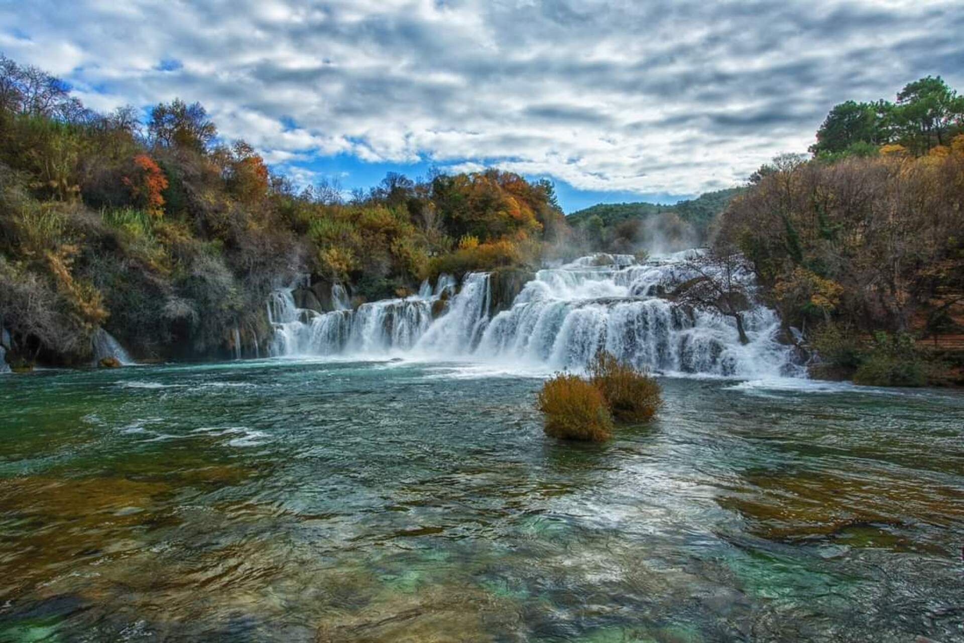 Ein Wasserfall stürzt in einen klaren Fluss, umgeben von Herbstlaub.
