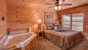 Quarto de cabana de madeira com uma cama grande, um ventilador de teto, uma janela com persianas e uma mesa de cabeceira de madeira com uma luminária.
