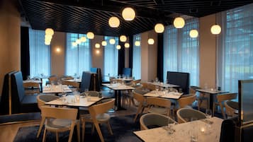 Ein Restaurant mit mehreren Tischen und Stühlen, großen Fenstern mit Vorhängen und hängenden, kugelförmigen Lampen.