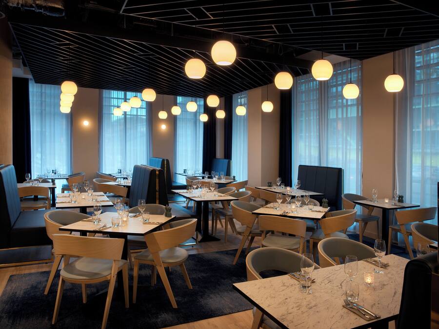 Ein Restaurant mit mehreren Tischen und Stühlen, großen Fenstern mit Vorhängen und hängenden, kugelförmigen Lampen.