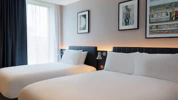 Habitación de hotel con dos camas, un ventanal con cortinas y cuadros enmarcados en la pared.
