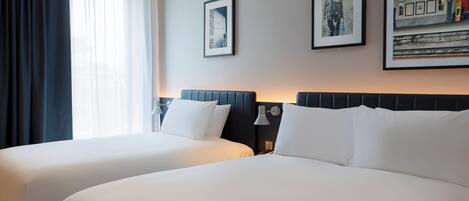 Habitación de hotel con dos camas, un ventanal con cortinas y cuadros enmarcados en la pared.