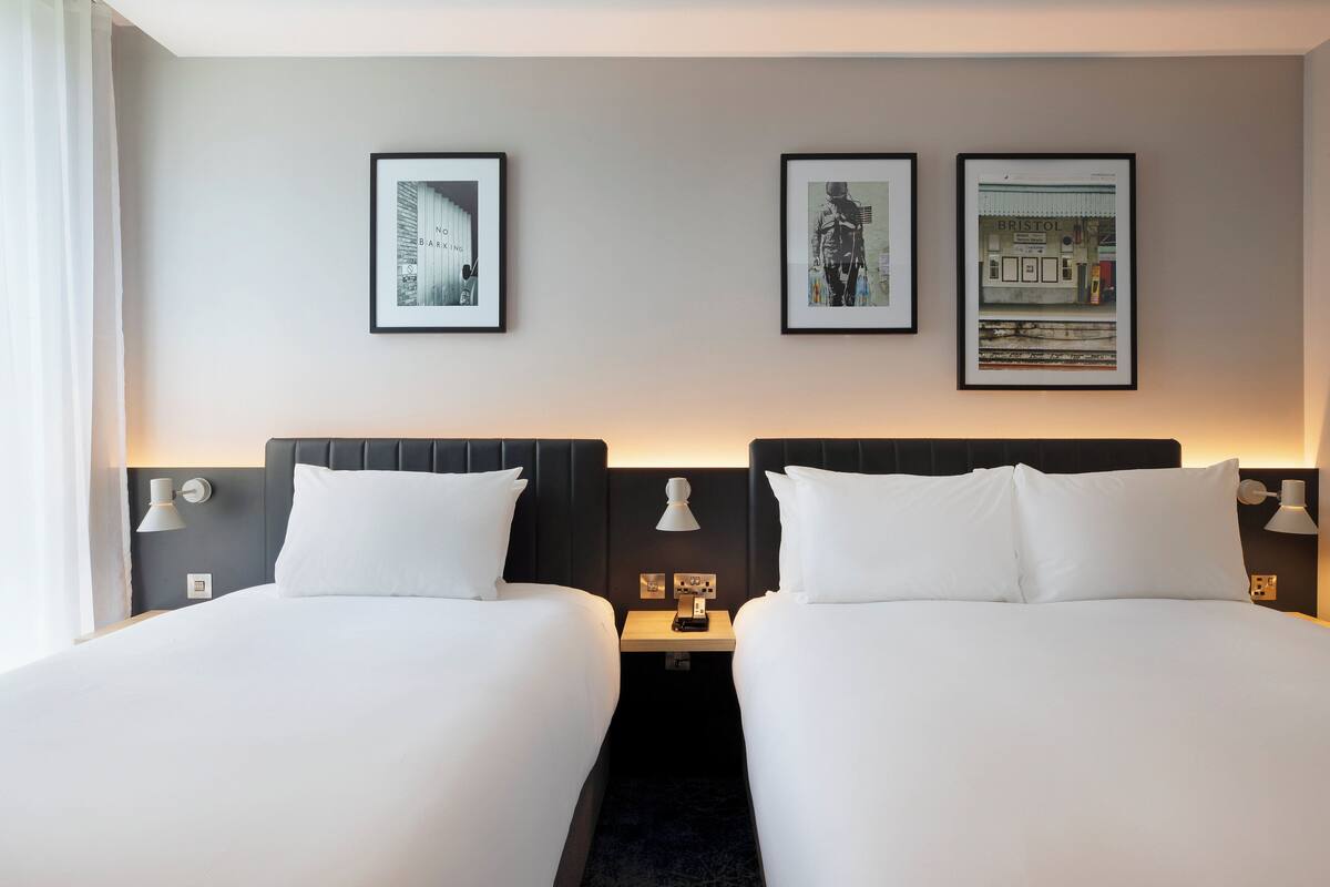 Quarto de hotel com duas camas, uma mesa de cabeceira com abajur, e três quadros na parede.