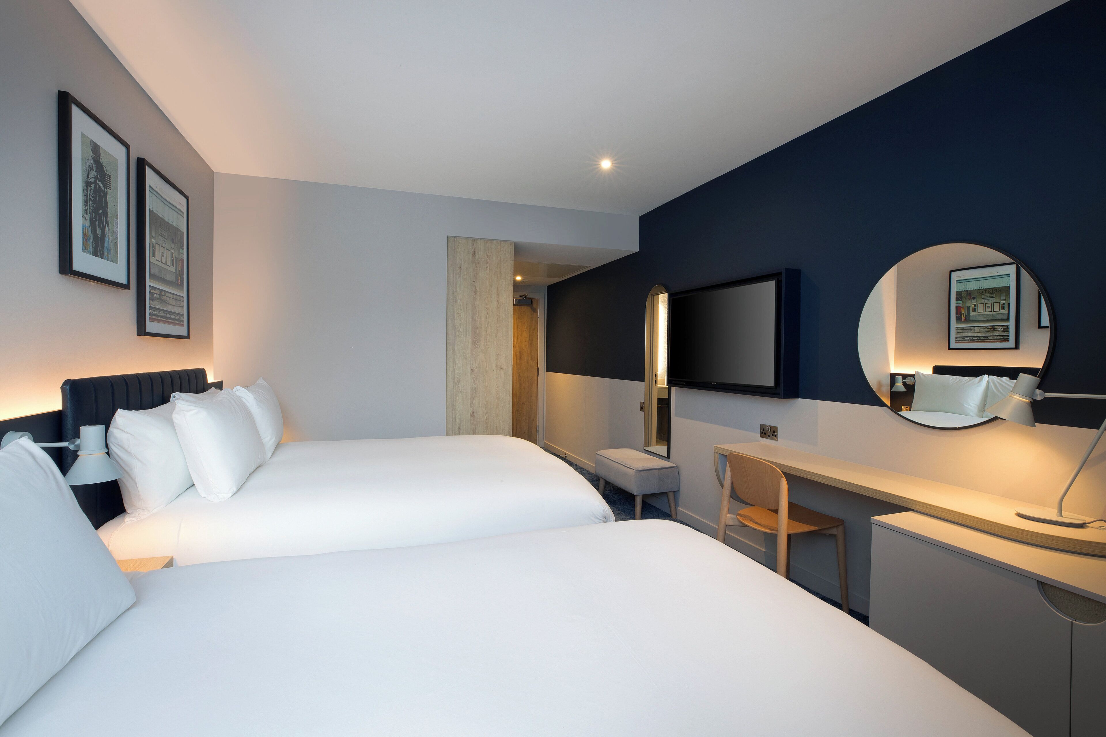 Habitación de hotel con dos camas, un escritorio, una silla, un espejo redondo y cuadros en la pared.