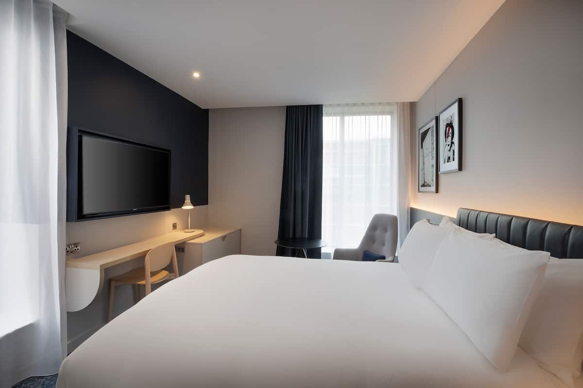 Quarto de hotel moderno com uma cama grande, uma escrivaninha, uma cadeira, uma televisão de tela plana e duas imagens emolduradas na parede.
