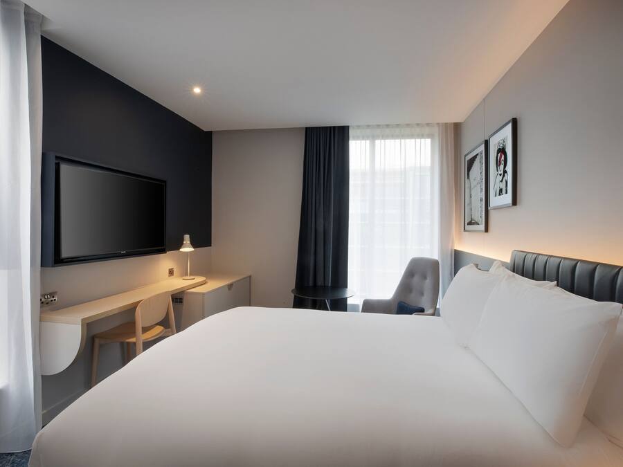 Une chambre d’hôtel moderne équipée d’un grand lit, d’un bureau, d’une chaise, d’un téléviseur à écran plat et de deux tableaux encadrés accrochés au mur.
