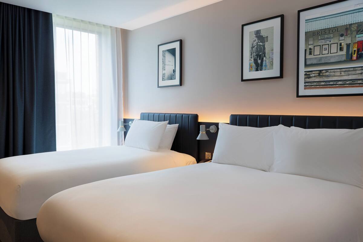 Quarto de hotel com duas camas, uma janela grande com cortinas e quadros nas paredes.