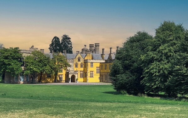 A large, historic building with a yellow facade, surrounded by lush green lawns and trees.