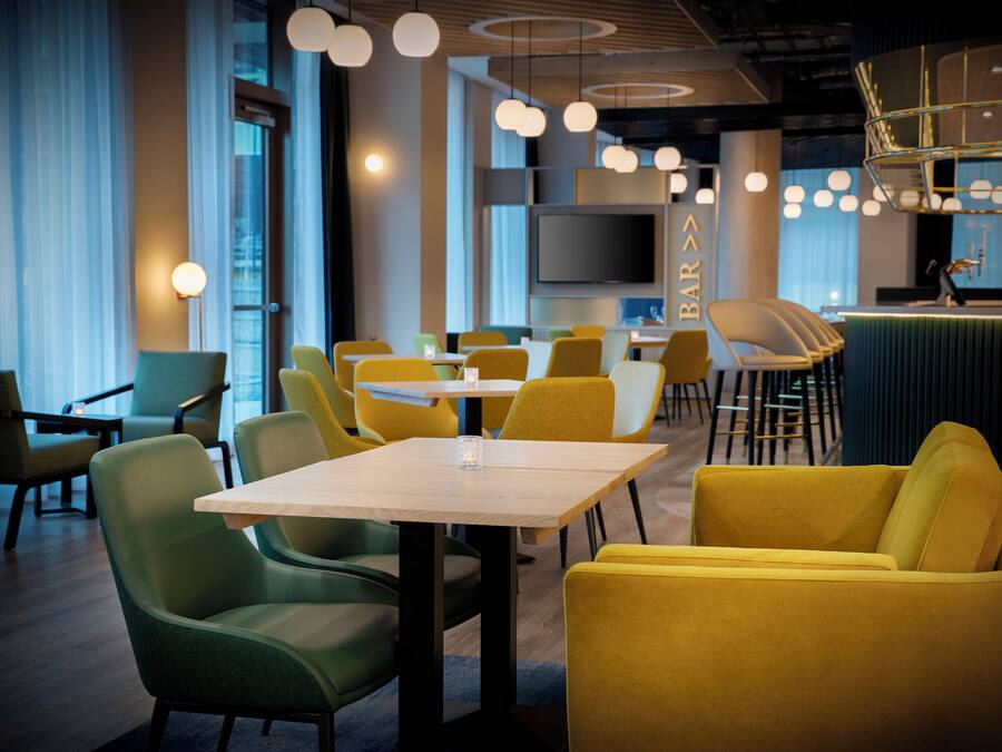 A modern restaurant with yellow and green chairs, a bar area, and a large TV screen.