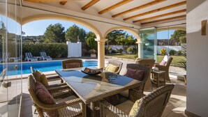 Alpendre coberto com móveis de vime, mesa de vidro e vista para uma piscina e jardim.