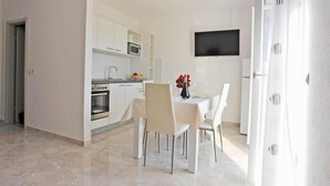 Une cuisine moderne avec des meubles blancs, un four à micro-ondes et un four traditionnel. On y trouve une table à manger avec des chaises et un vase de fleurs. Un téléviseur à écran plat est fixé au mur, au-dessus de la table.