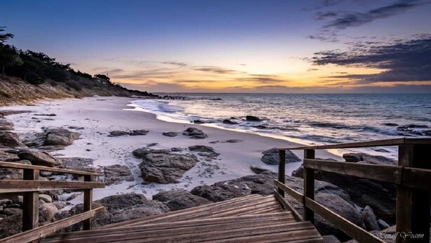Une promenade en bois menant à une plage au terrain rocheux, avec un coucher de soleil en arrière-plan.