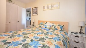 Ein Schlafzimmer mit einem blumigen Bettüberwurf, einem Holzkopfstück, einem Nachttisch mit Lampe, einem Beistelltisch und einem Wandschild mit der Aufschrift 'BEACH'.