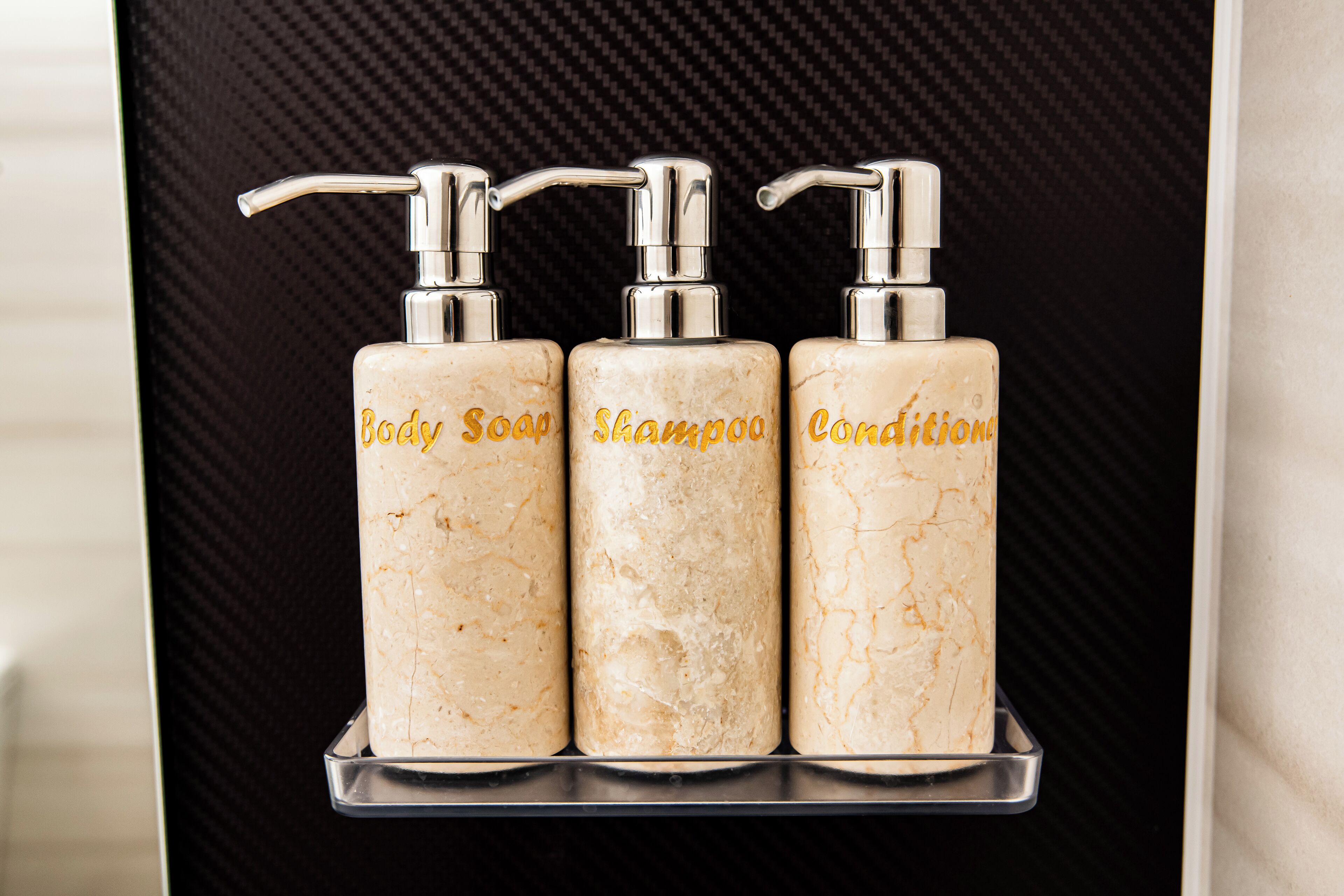 Tres dispensadores de baño etiquetados como "Body Soap", "Shampoo" y "Conditioner" sobre una encimera negra.