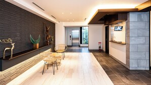 Een moderne lobby met een houten receptiebalie, een zithoek met rieten stoelen, een wandtelevisie en uitzicht op de lift.