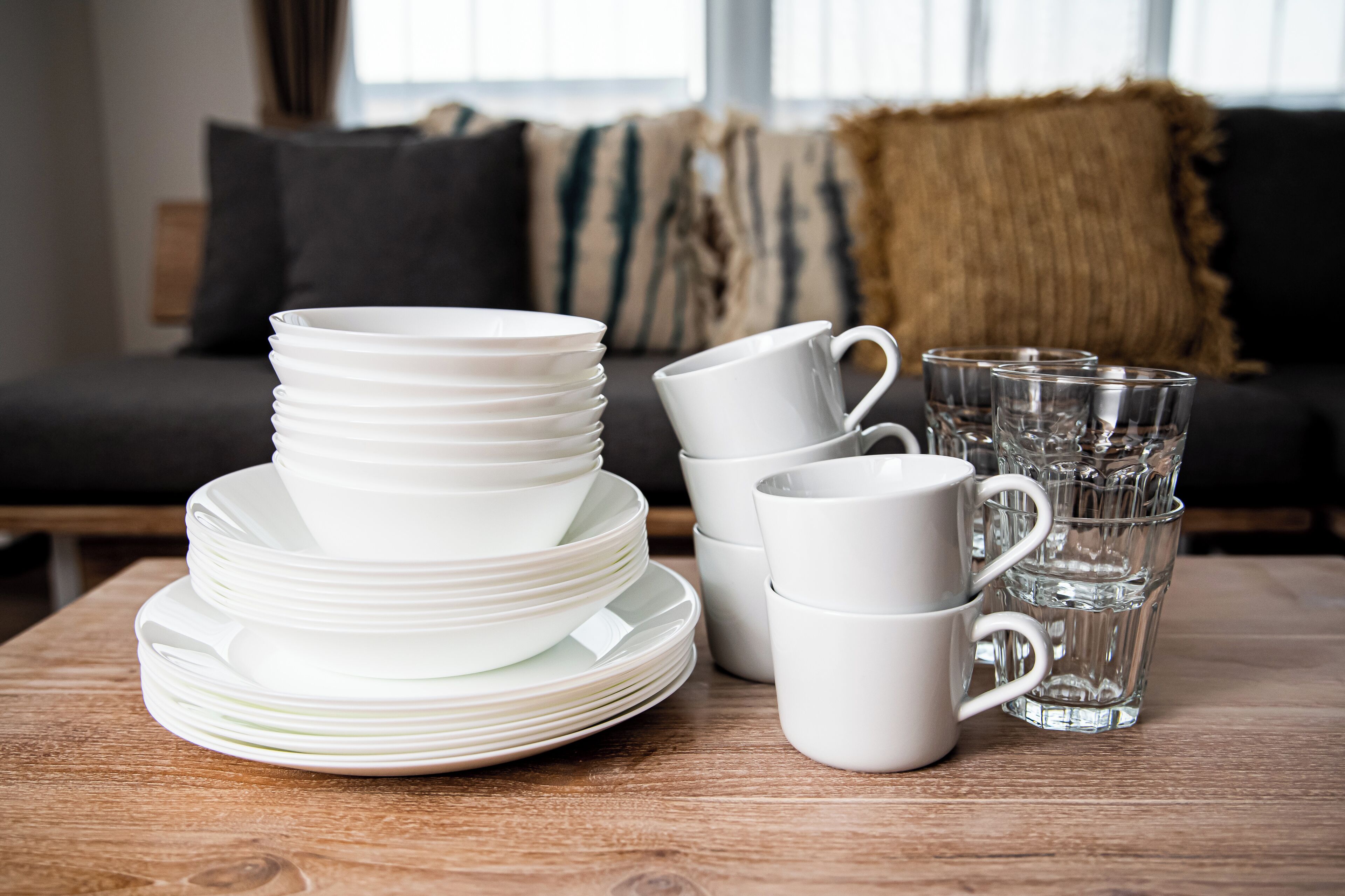 A stack of white plates and cups on a wooden table, with a glass of water and a sofa in the background.