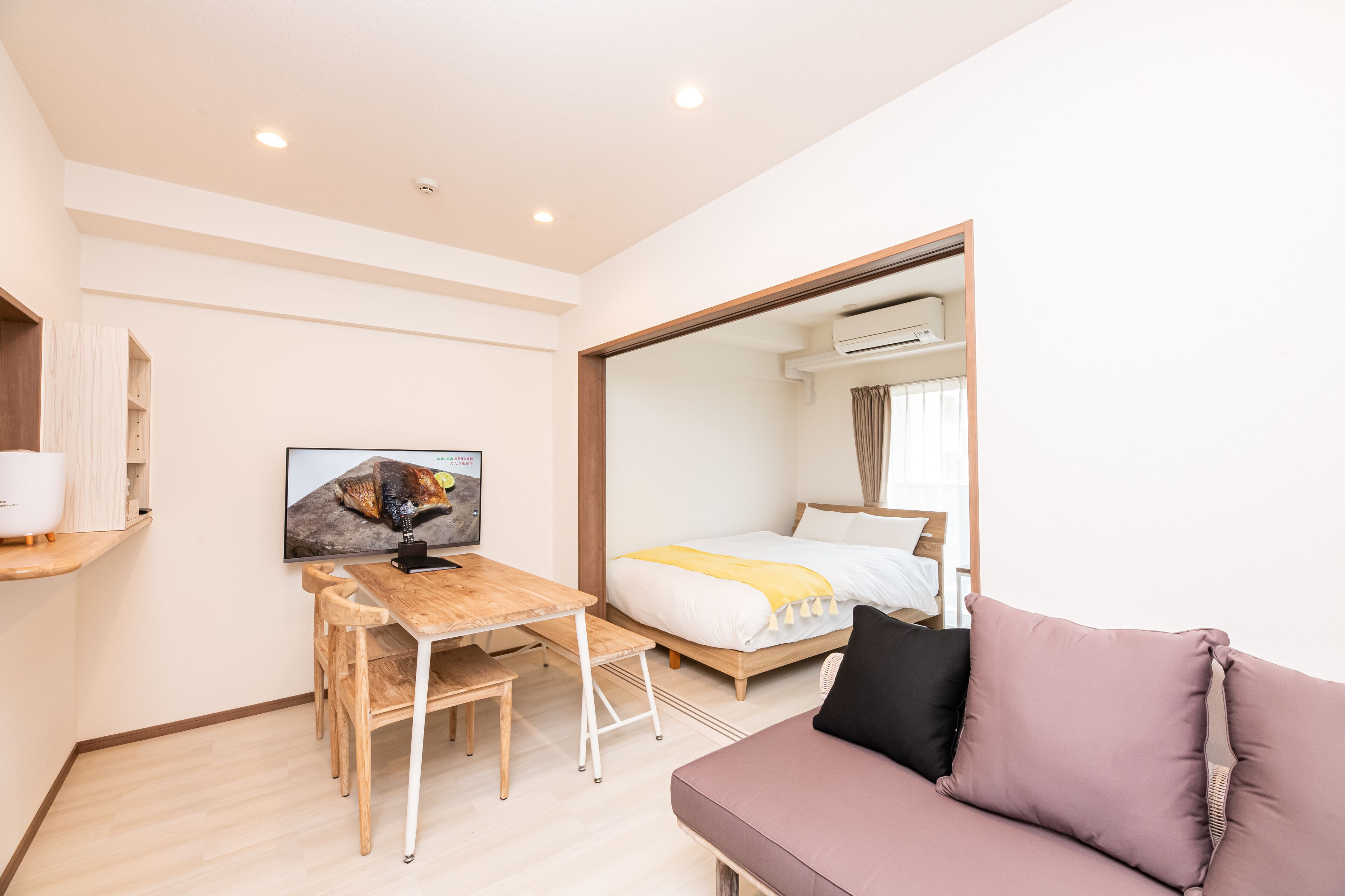 Une chambre d’hôtel moderne avec un bureau en bois, un lit recouvert d’une courtepointe blanche et jaune, un grand miroir et un canapé violet.