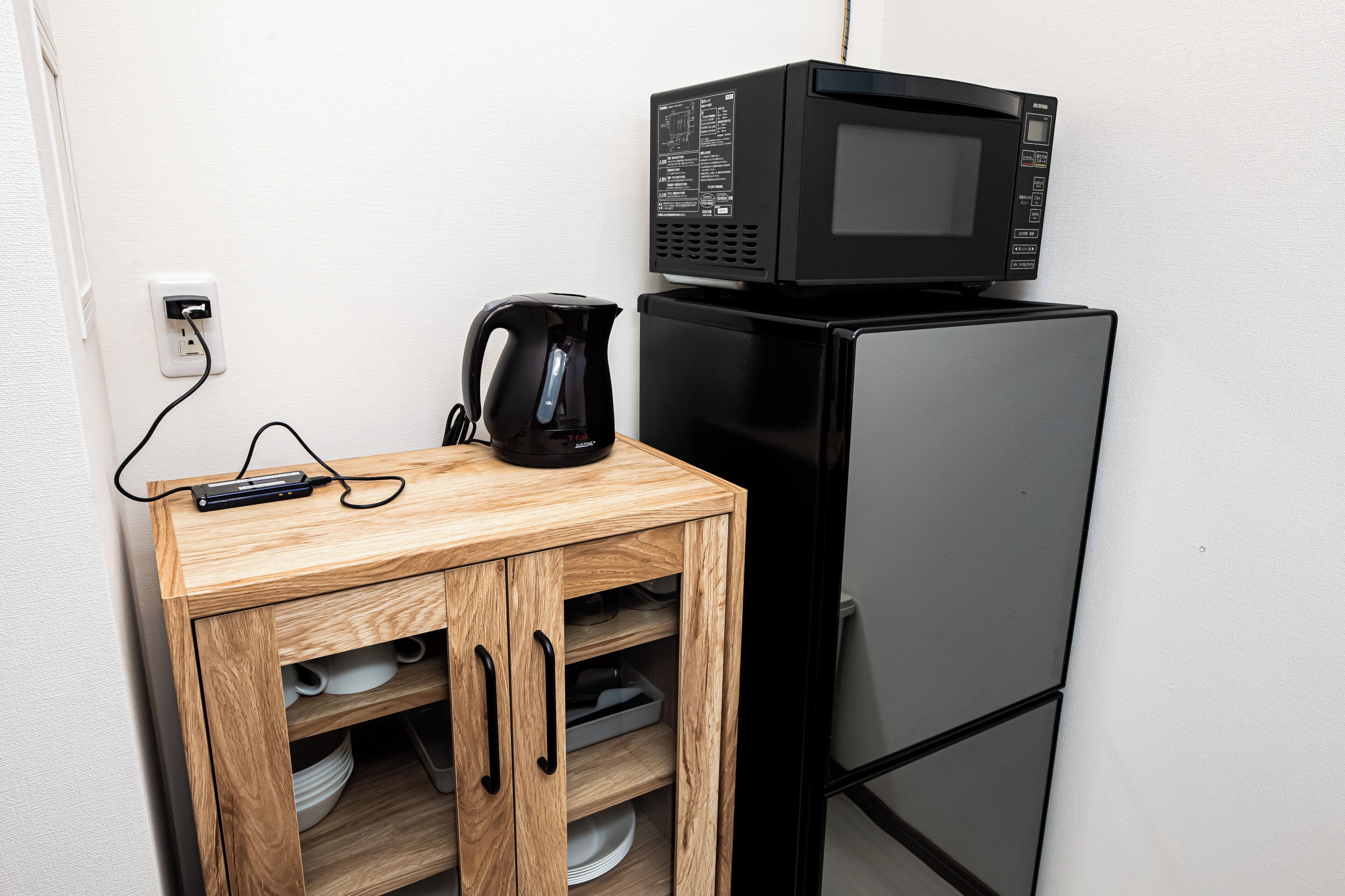 Uma kitchenette compacta com um frigorífico preto, um micro-ondas, um armário de madeira com prateleiras e uma chaleira.