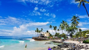 Una playa con aguas turquesas cristalinas, palmeras y un edificio con balcón con vista al océano.