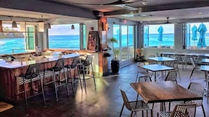 Un restaurante con zona de bar, mesas al aire libre y grandes ventanales con vistas al mar.