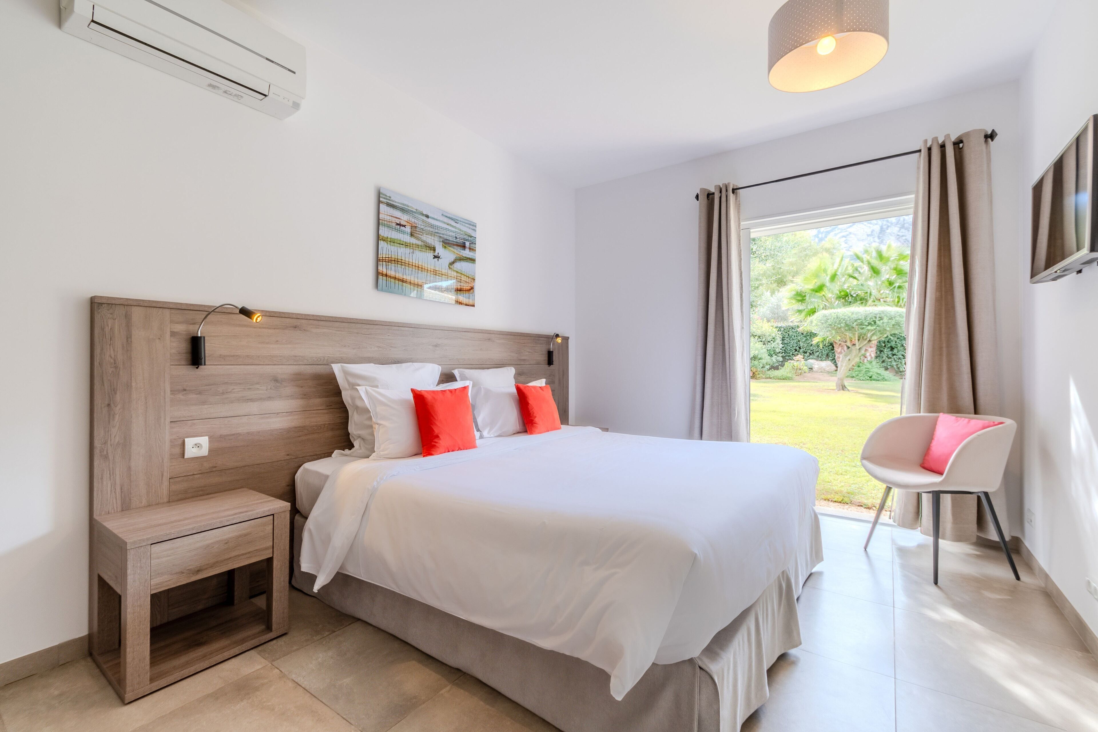A bedroom with a wooden headboard, white bedding, a small bedside table, a chair, and a view of the garden through the window.