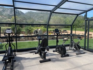 A gym with various exercise equipment, including treadmills and stationary bikes, with a view of mountains and greenery through large windows. - Exclusive Four-Bedroom Villa featuring a Private Pool, Garden, Parking and Spa (CALVI)
