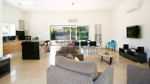 A modern living room with a sofa, coffee table, and TV. There is a kitchen area with a sink and cabinets. - Exclusive Four-Bedroom Villa featuring a Private Pool, Garden, Parking and Spa (CALVI)