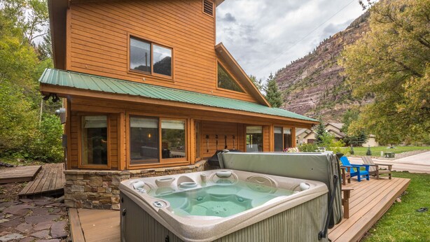 Uma cabana de madeira com banheira de hidromassagem em um deck, rodeada por um muro de pedra e um panorama de montanhas.