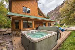 A wooden cabin with a hot tub on a deck, surrounded by a stone wall and a mountain backdrop.
