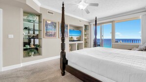A bedroom with a large bed, a ceiling fan, a TV, a bookshelf, and a balcony with a view.
