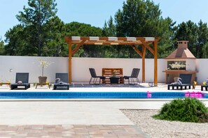 Un área de piscina con sillones de descanso, una pérgola de madera y una parrilla.