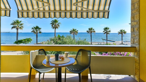A balcony with a striped awning, two chairs, a small round table, and a view of the sea and palm trees. - Beachfront Holiday Apartment Inga Yellow with sea view (Vir)