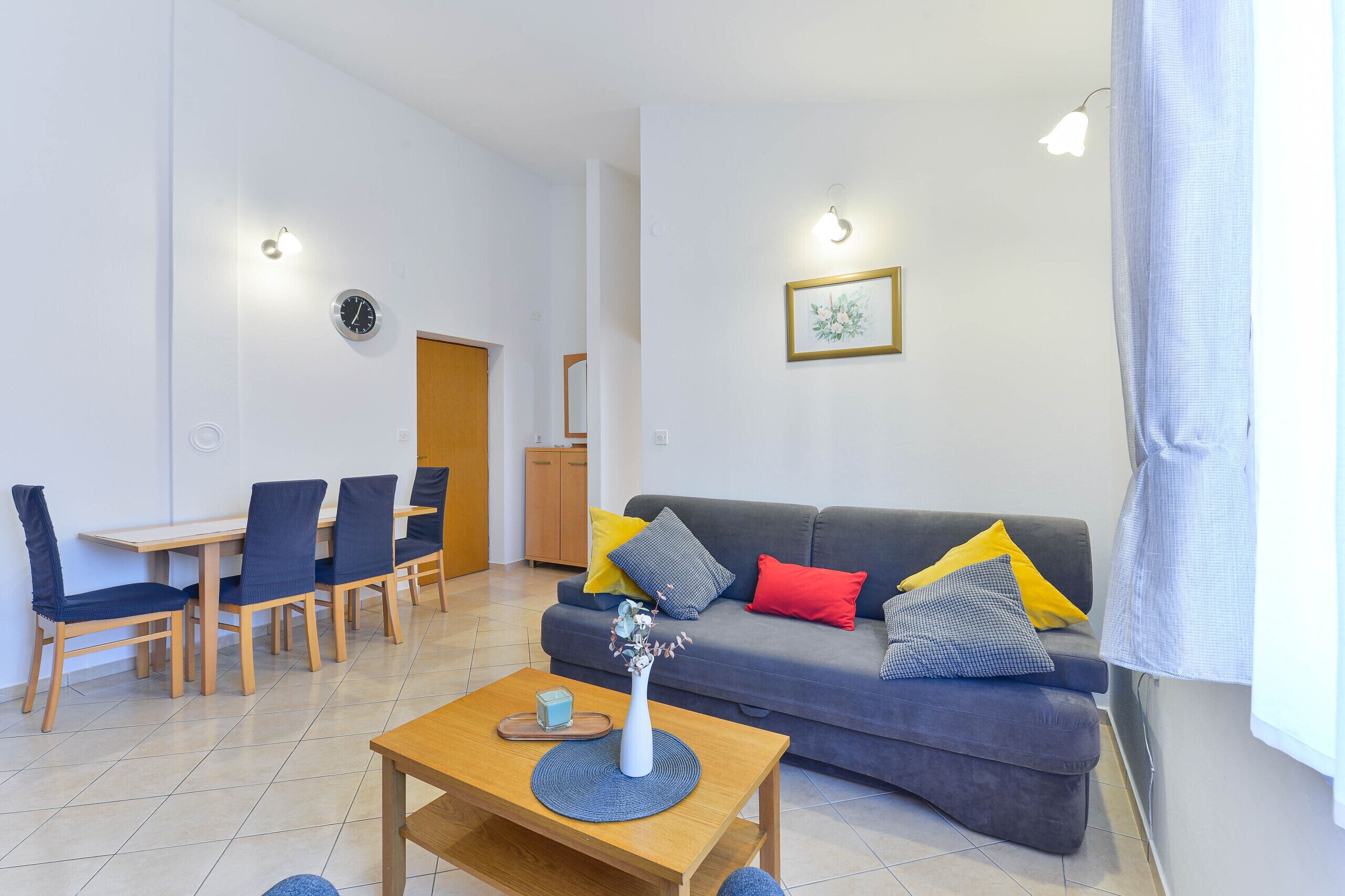 Un soggiorno moderno con un divano grigio, cuscini blu e gialli, un tavolino in legno e un'area pranzo con sedie blu e un tavolo in legno.