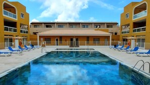 A swimming pool with blue lounge chairs and a building with yellow walls and a tiled roof.