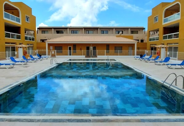 A swimming pool with blue lounge chairs and a building with yellow walls and a tiled roof.