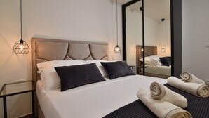 Een net opgemaakt bed met hoofdbord, twee bedlampjes, een spiegel en opgerolde handdoeken op het bed.