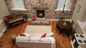 A cozy living room with a stone fireplace, a piano, a sofa, a dining table, and a kitchen area.