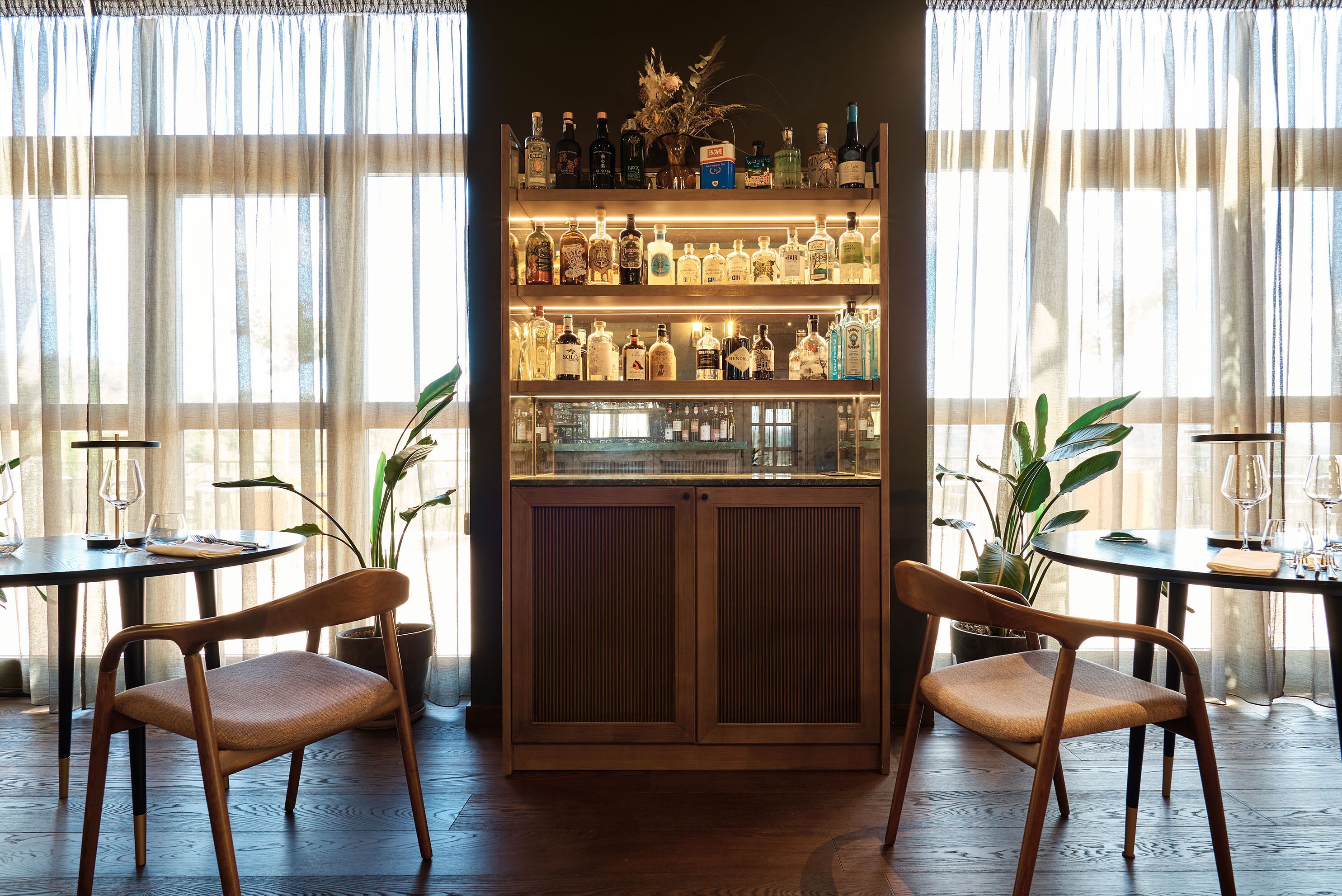 A bar area with wooden chairs, a liquor display, and large windows with sheer curtains.
