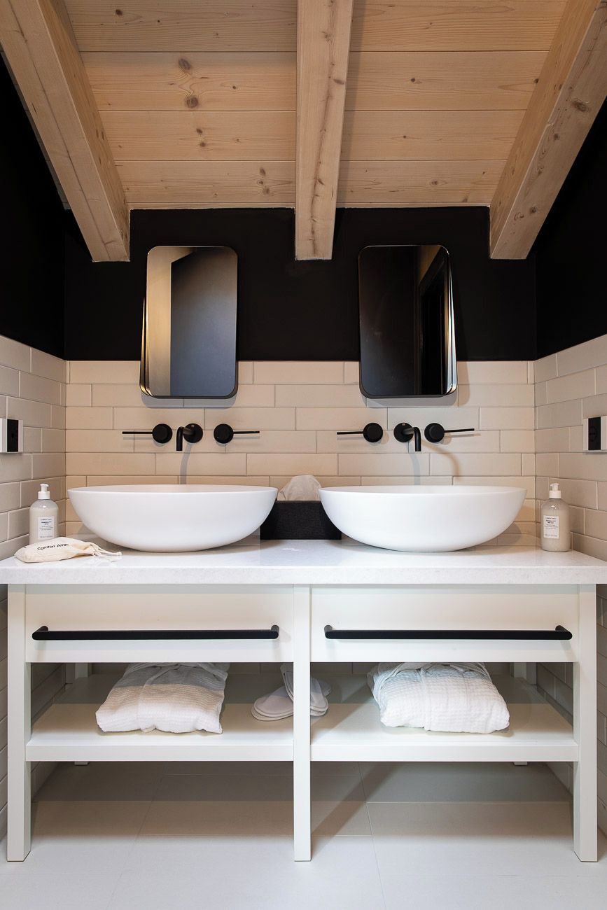 Un baño moderno con dos lavamanos blancos, grifos negros y techo de madera.
