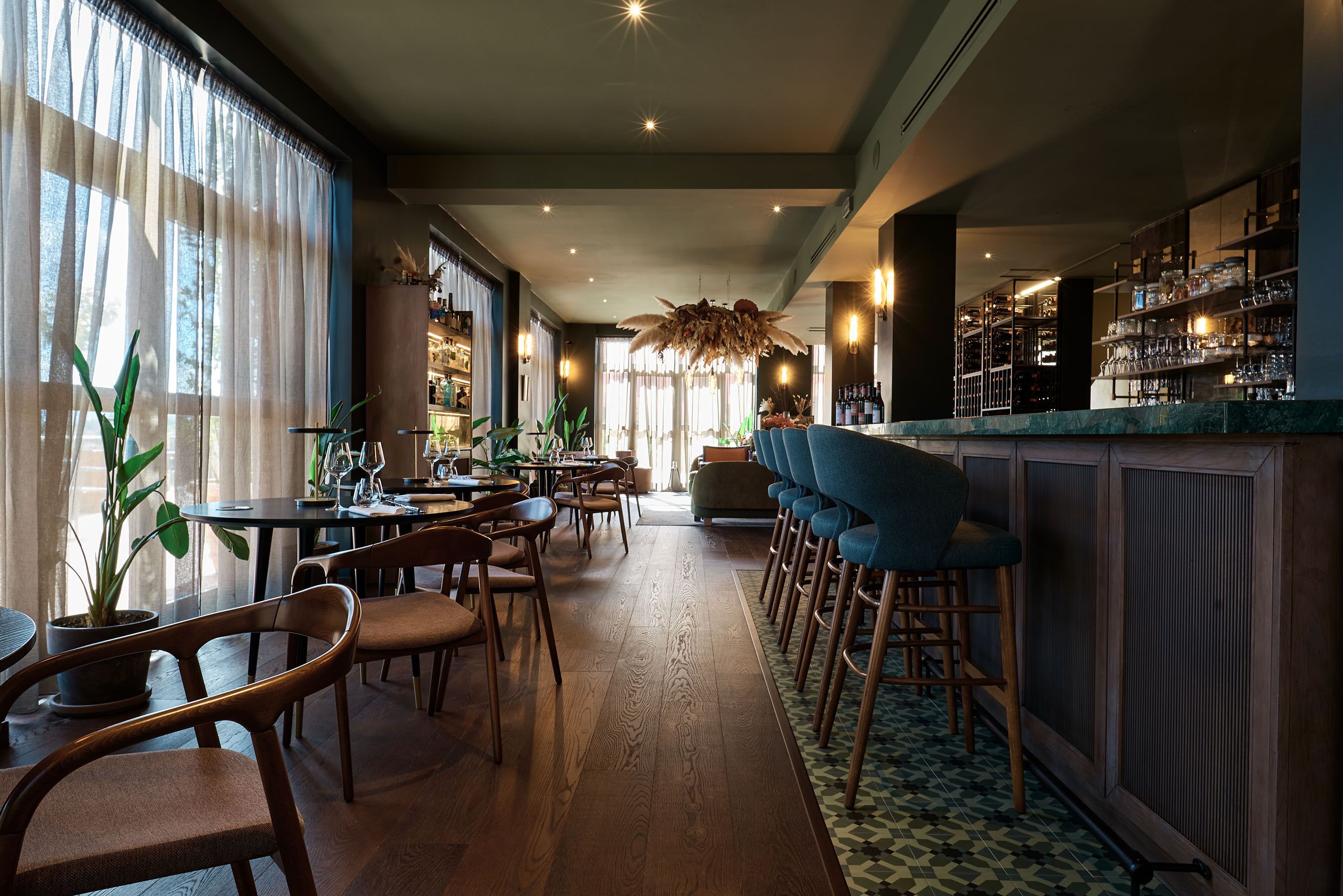 Zona de bar con sillas y mesas de madera, barra con estantes llenos de botellas y un ventanal con cortinas traslúcidas.